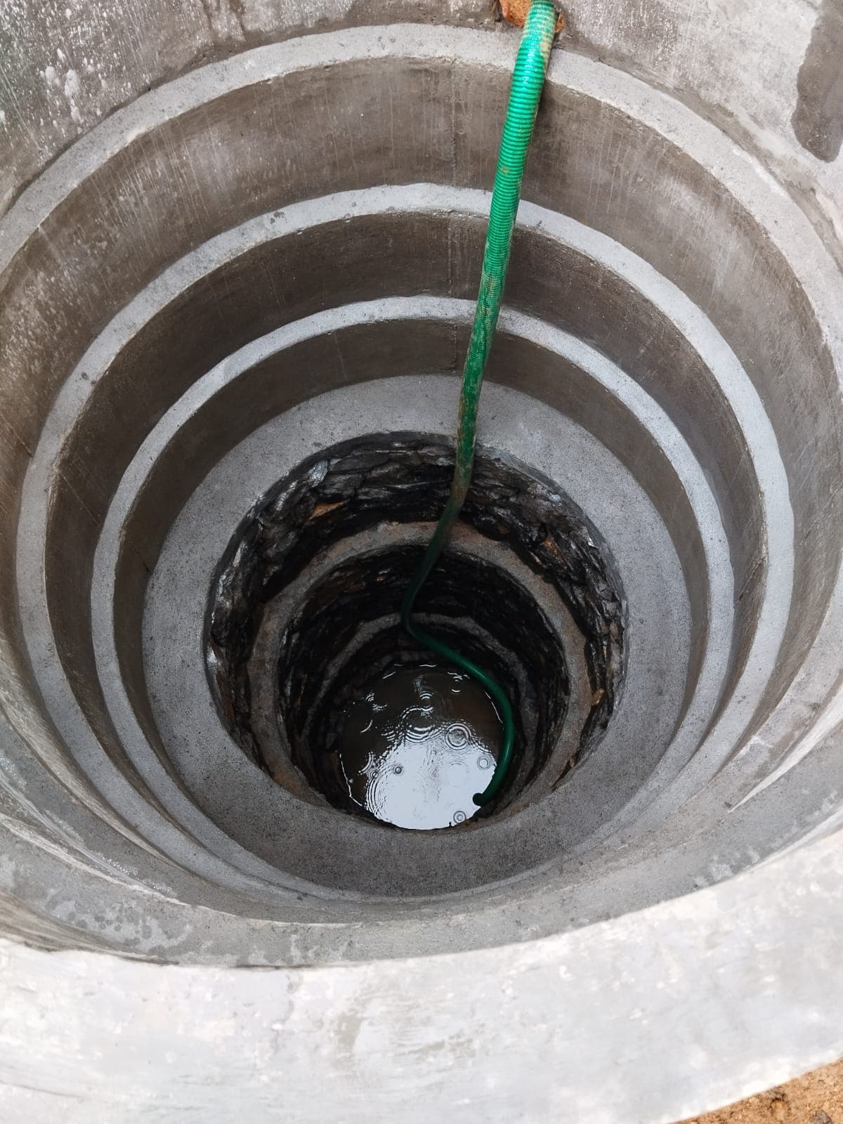 Deep circular well lined with concrete rings and stones, with a green hose reaching water.