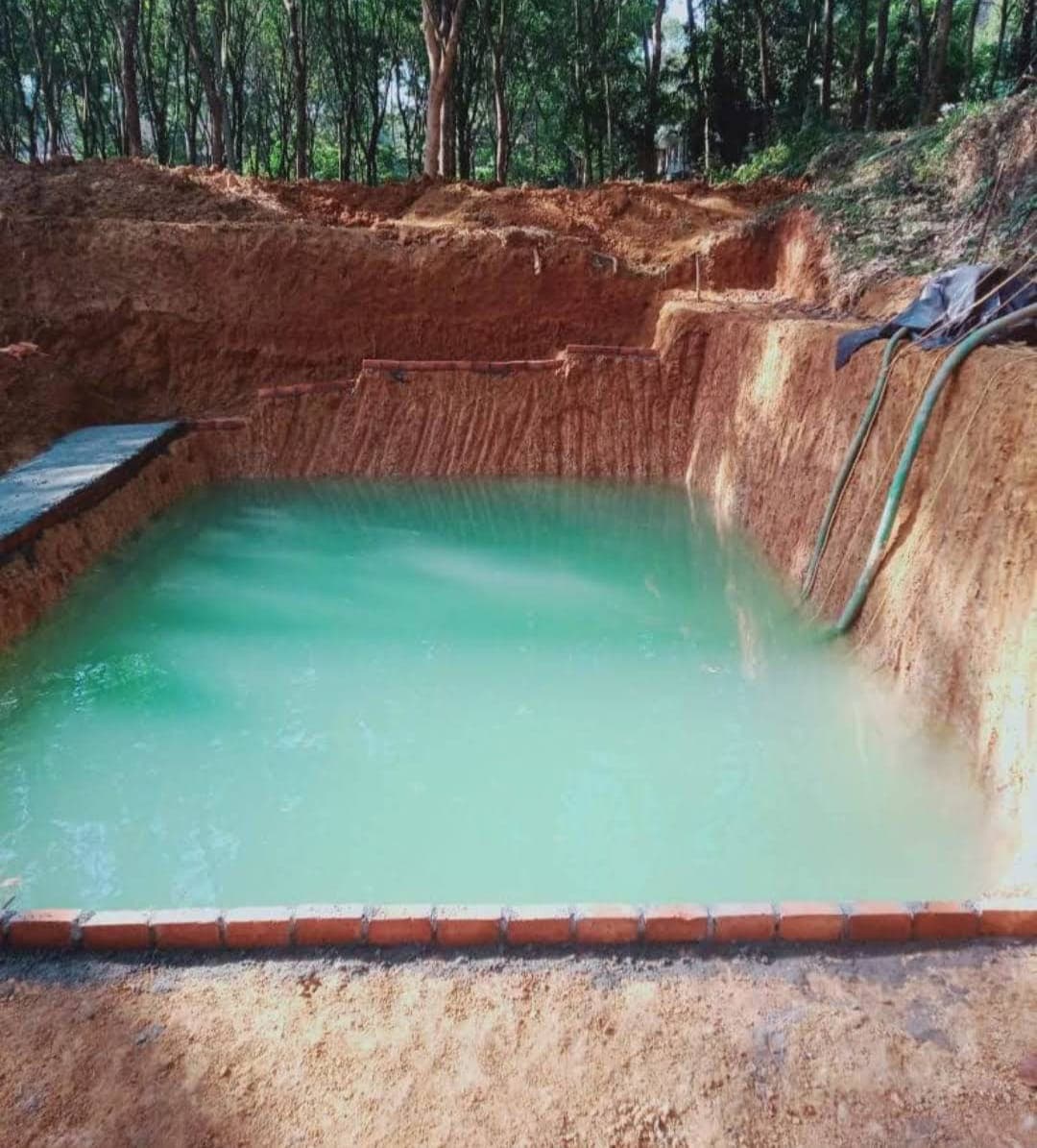 Rectangular earthen pit filled with turquoise water, surrounded by red clay and dense forest.