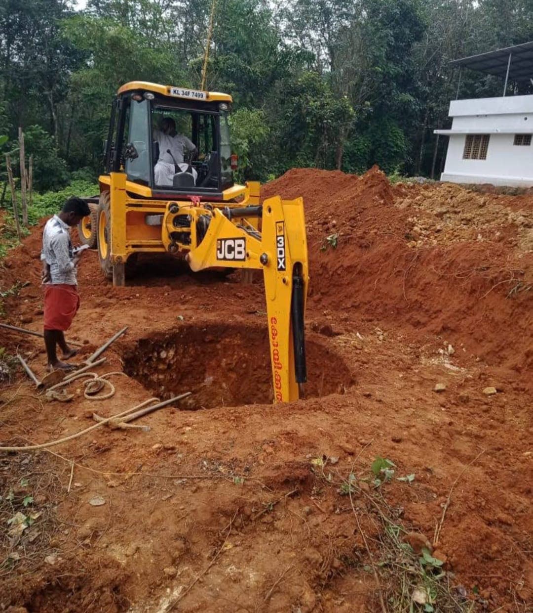 Yellow JCB backhoe digging a deep circular pit in red soil at a construction site.