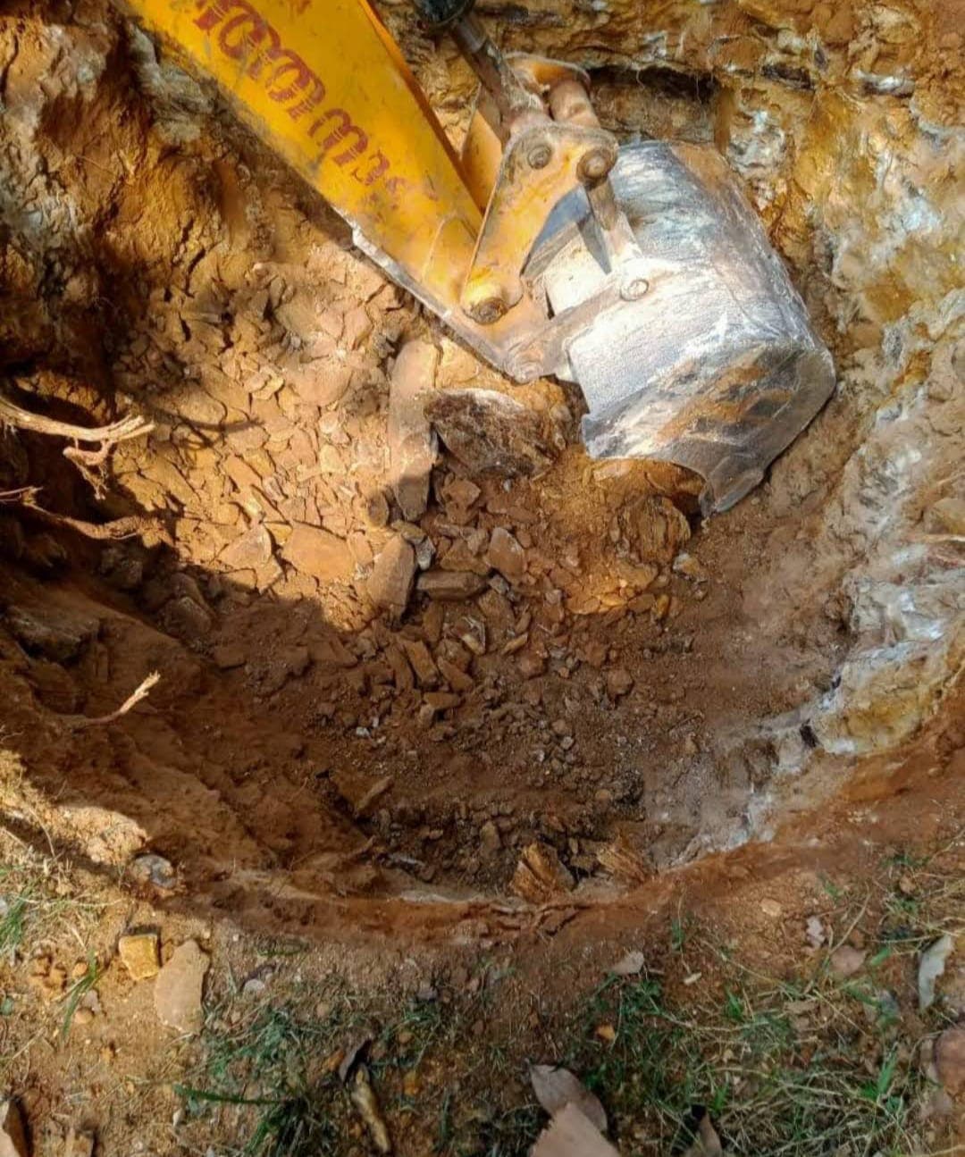 Yellow excavator bucket digging a deep, circular hole into rocky, reddish-brown soil and earth.