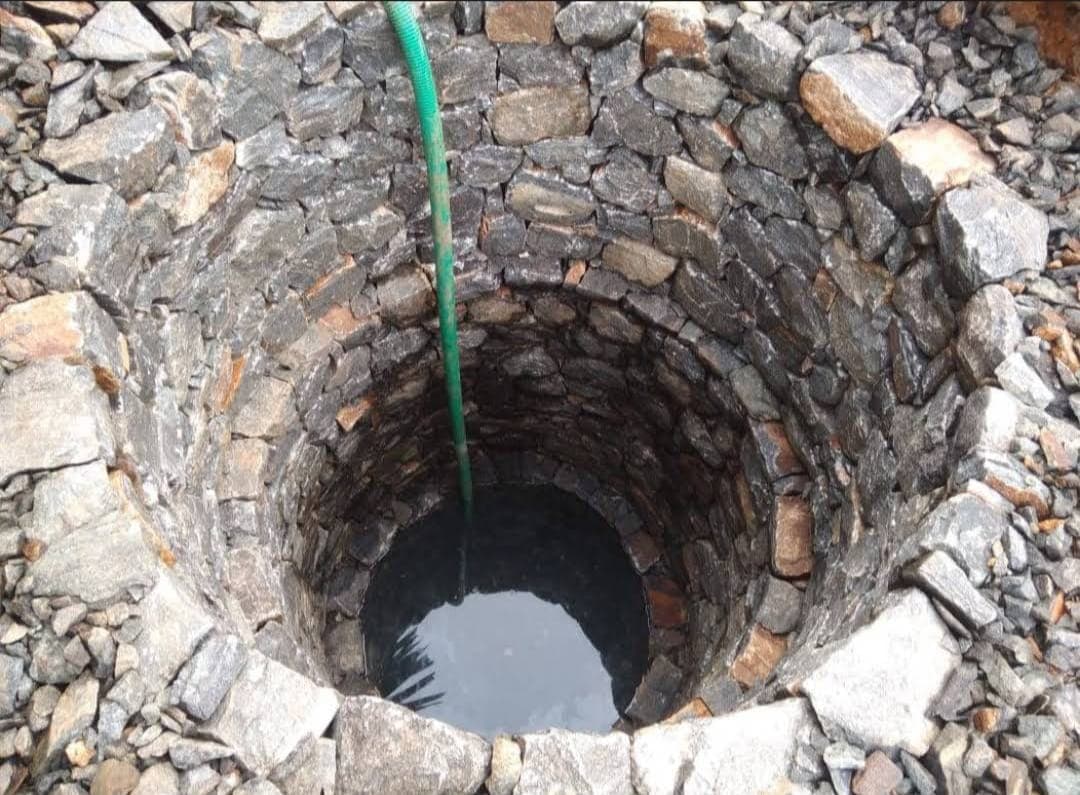 Deep stone-lined well with dark water at the bottom and a green hose descending.