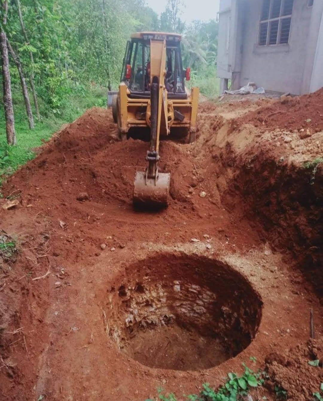 Yellow backhoe digging a deep circular hole in red soil next to a building.