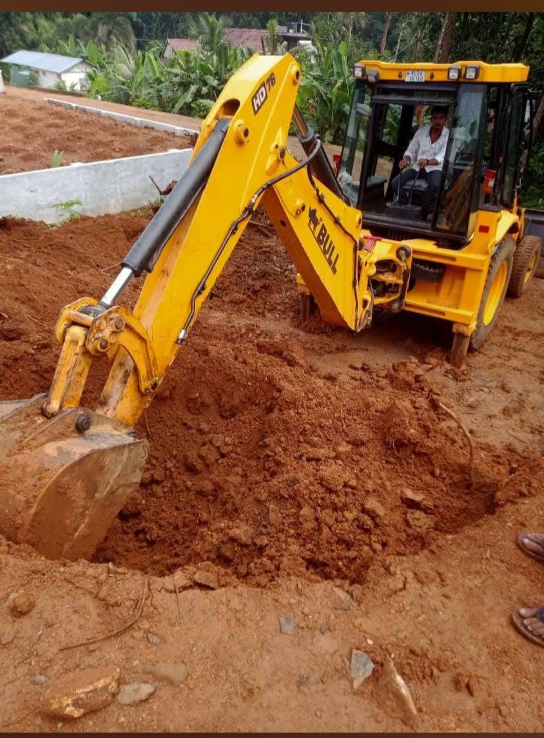 Yellow backhoe loader digging a deep hole in red soil at a construction site.